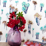 A vase of flowers stands in front of a wall papered in a playful palm tree print in shades of brown, gray and green on a white field.