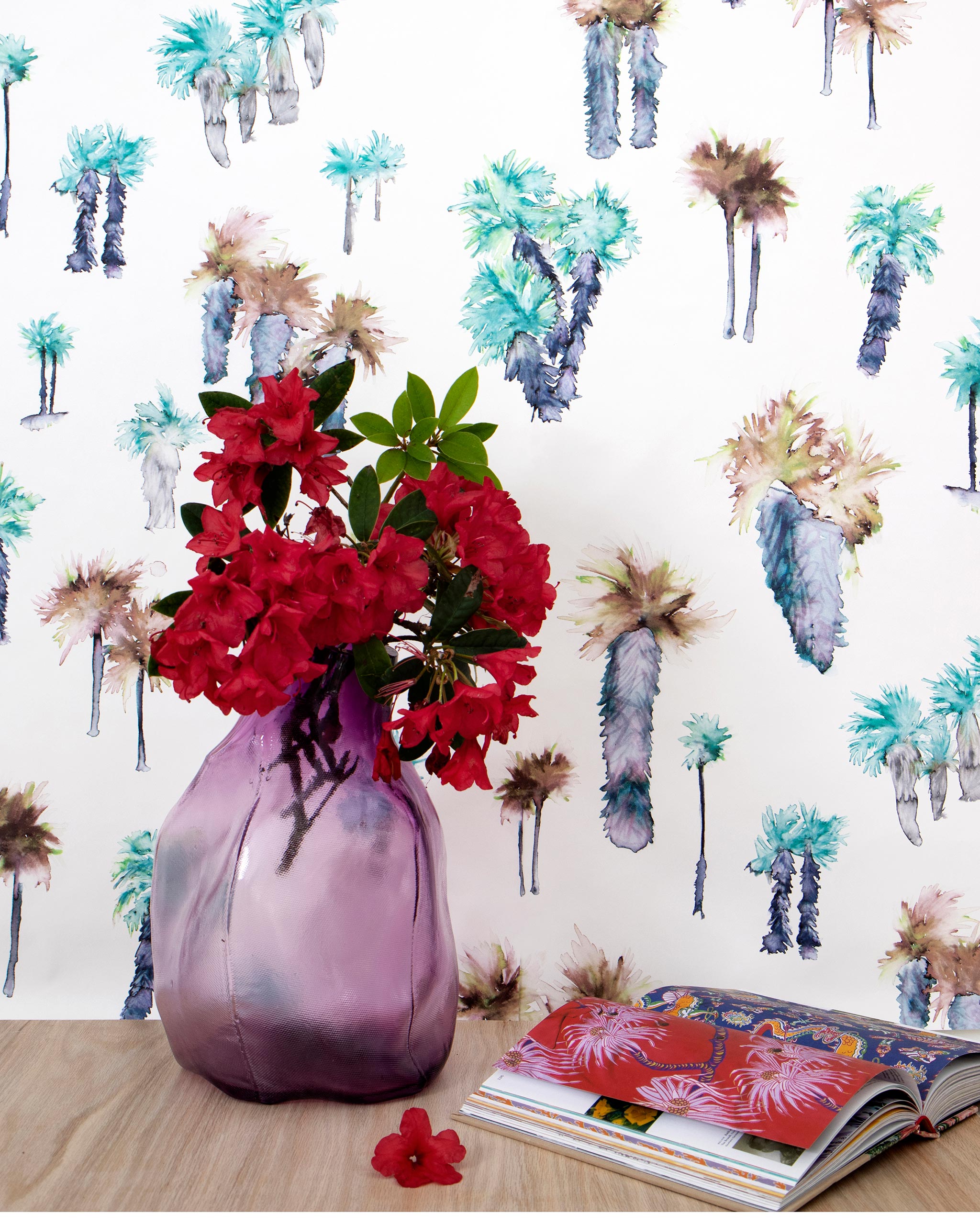 A vase of flowers stands in front of a wall papered in a playful palm tree print in shades of brown, gray and green on a white field.