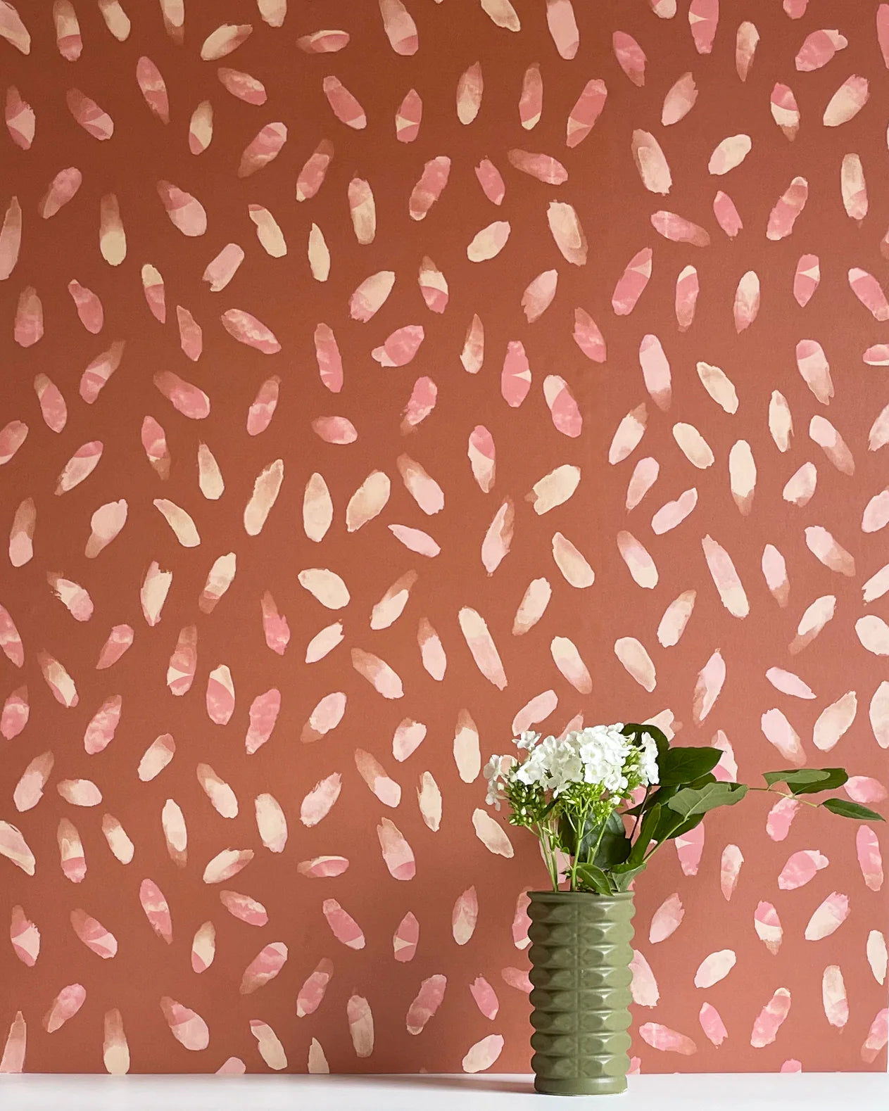 A vase of flowers stands in front of a wall covered in a playful paint blotch print in pink and cream on a brown field.