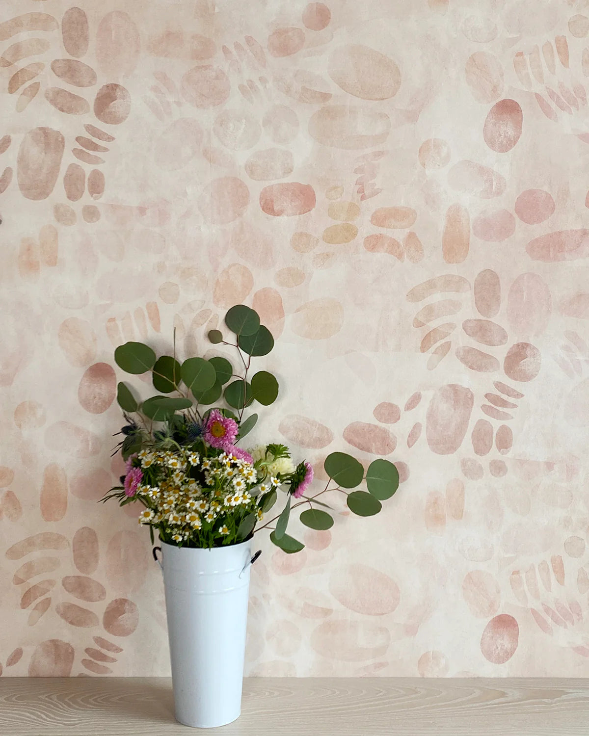 A vase of flowers stands in front of a wall covered in a playful paint blotch print in light pink on a cream field.