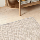 Herringbone patterned area rug in taupe on a wood floor with a bookshelf. 