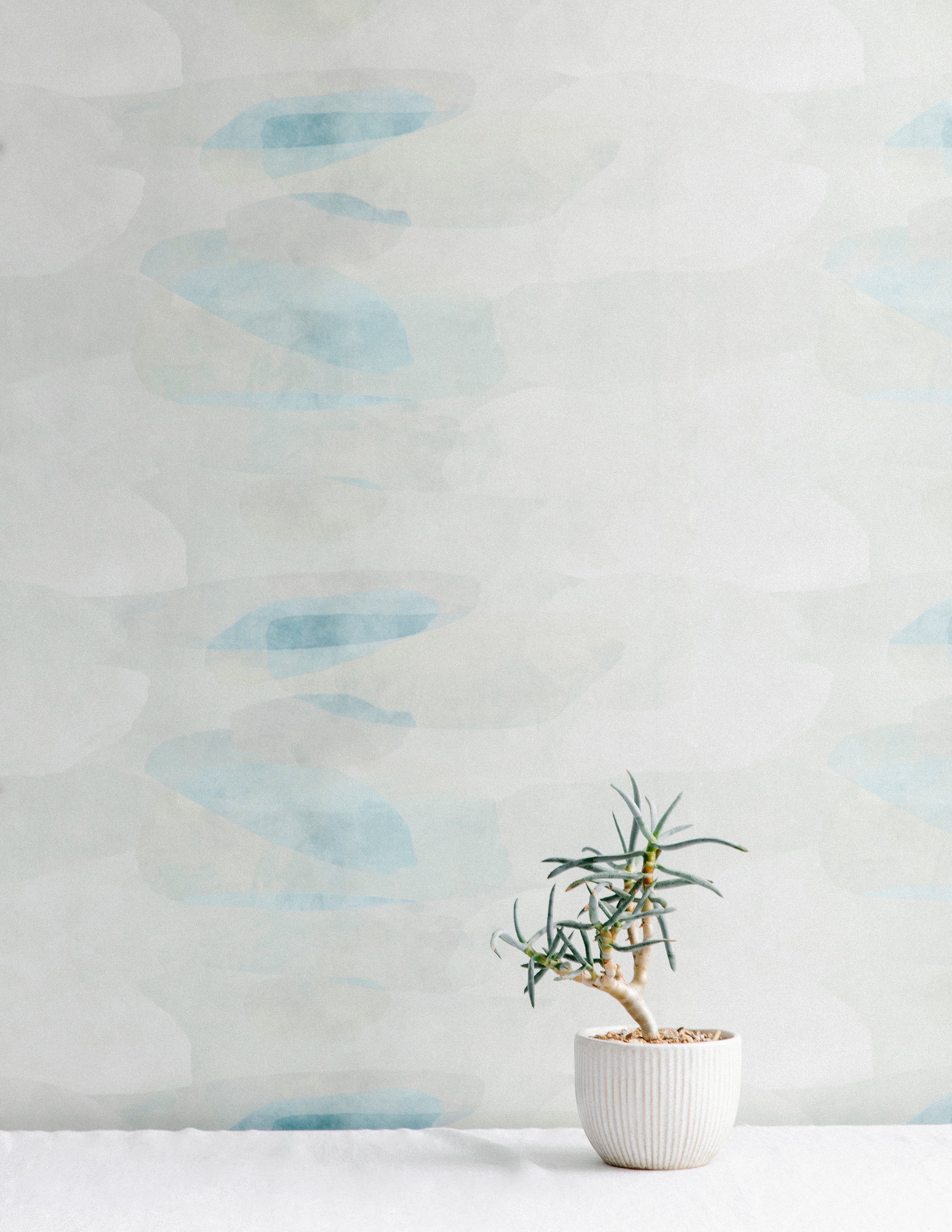 A potted plant stands in front of a wall covered in an abstract color blot print in shades of white, gray and turquoise.