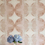 A vase of flowers stands in front of a wall covered in a cloudy ink blot print in shades of brown and pink on a cream field.