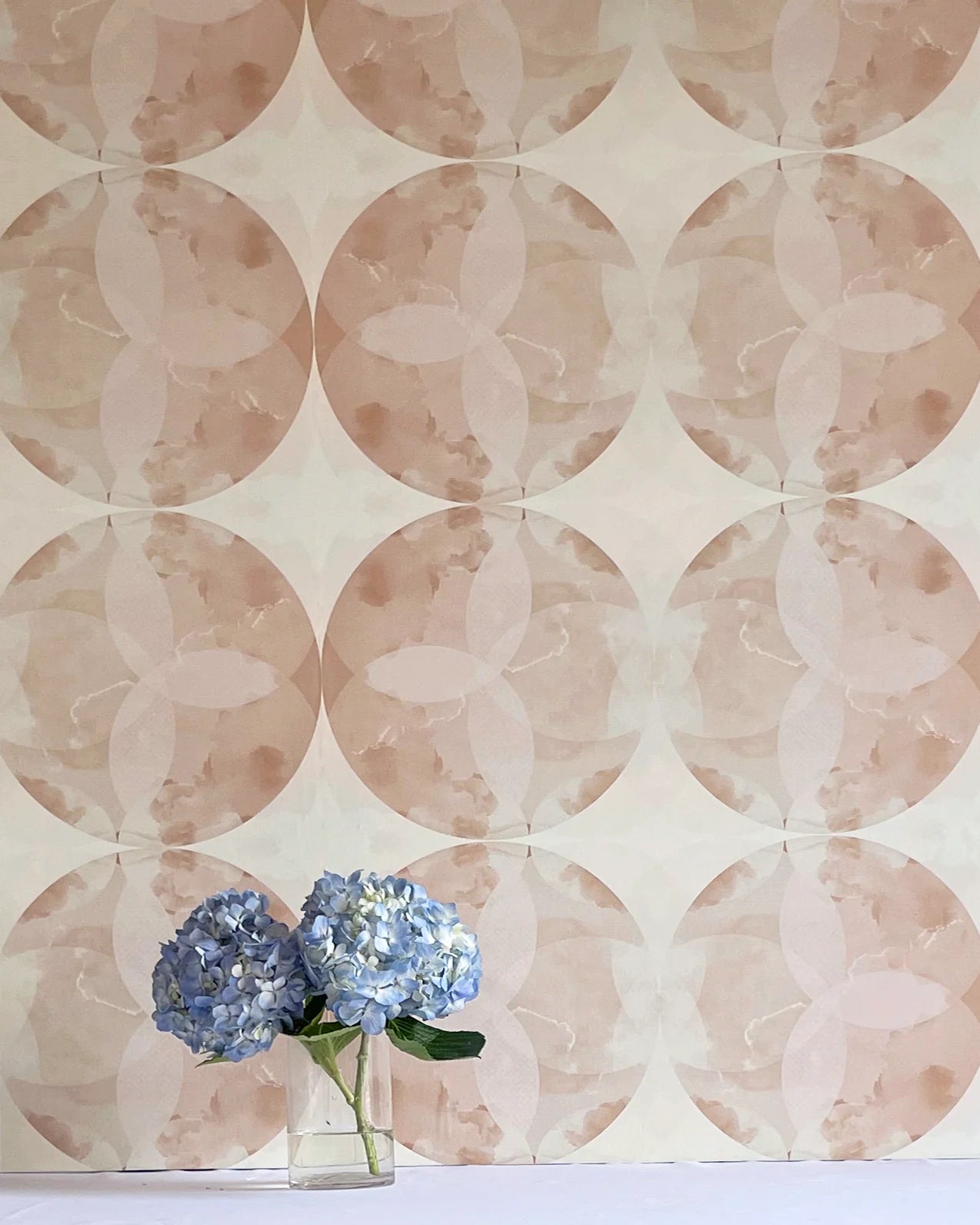 A vase of flowers stands in front of a wall covered in a cloudy ink blot print in shades of brown and pink on a cream field.