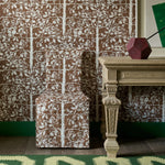 Slip cover chair in brown tree pattern fabric against matching wallpaper with an ornate wooden table and green rug. 