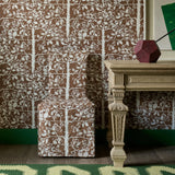 Slip cover chair in brown tree pattern fabric against matching wallpaper with an ornate wooden table and green rug. 