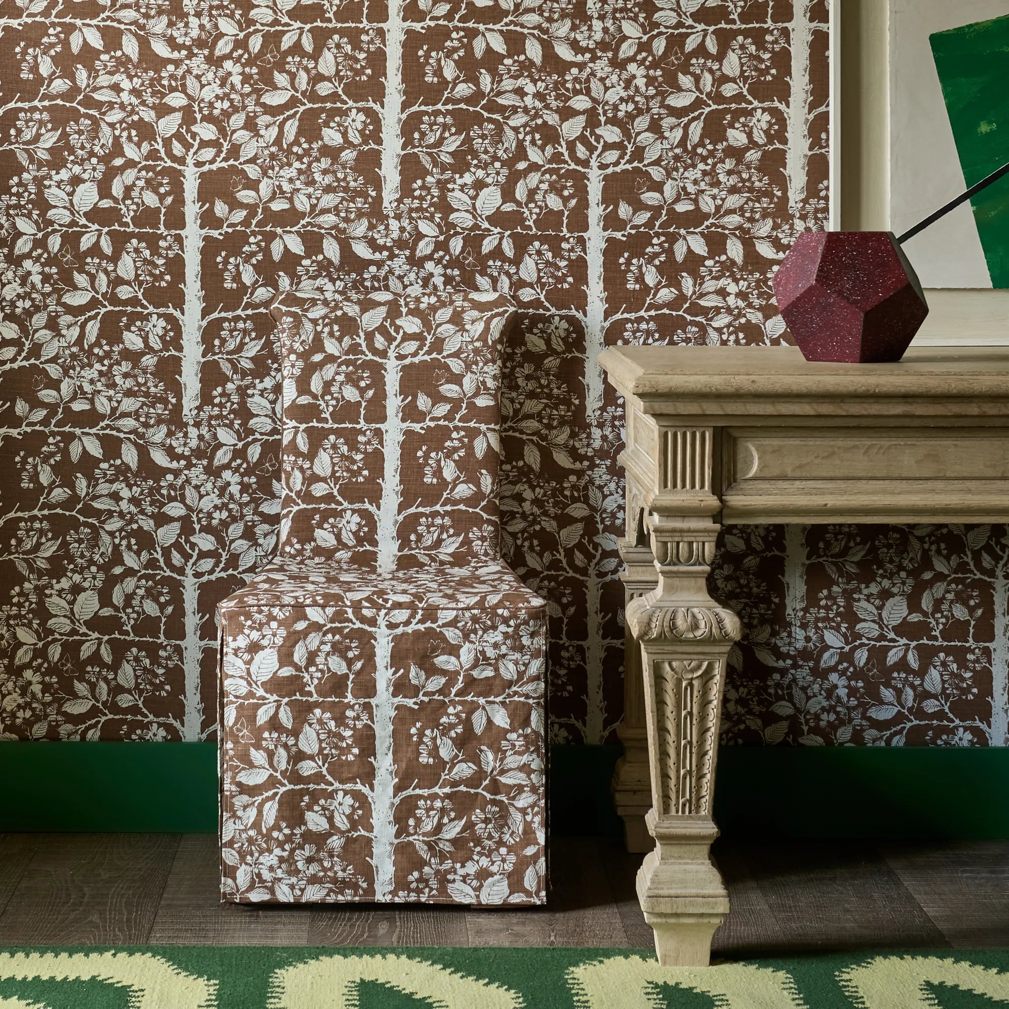 Slip cover chair in brown tree pattern fabric against matching wallpaper with an ornate wooden table and green rug. 