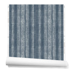 A partially unrolled roll of wallpaper in a striped pattern of intricate white lines over a washed denim background.