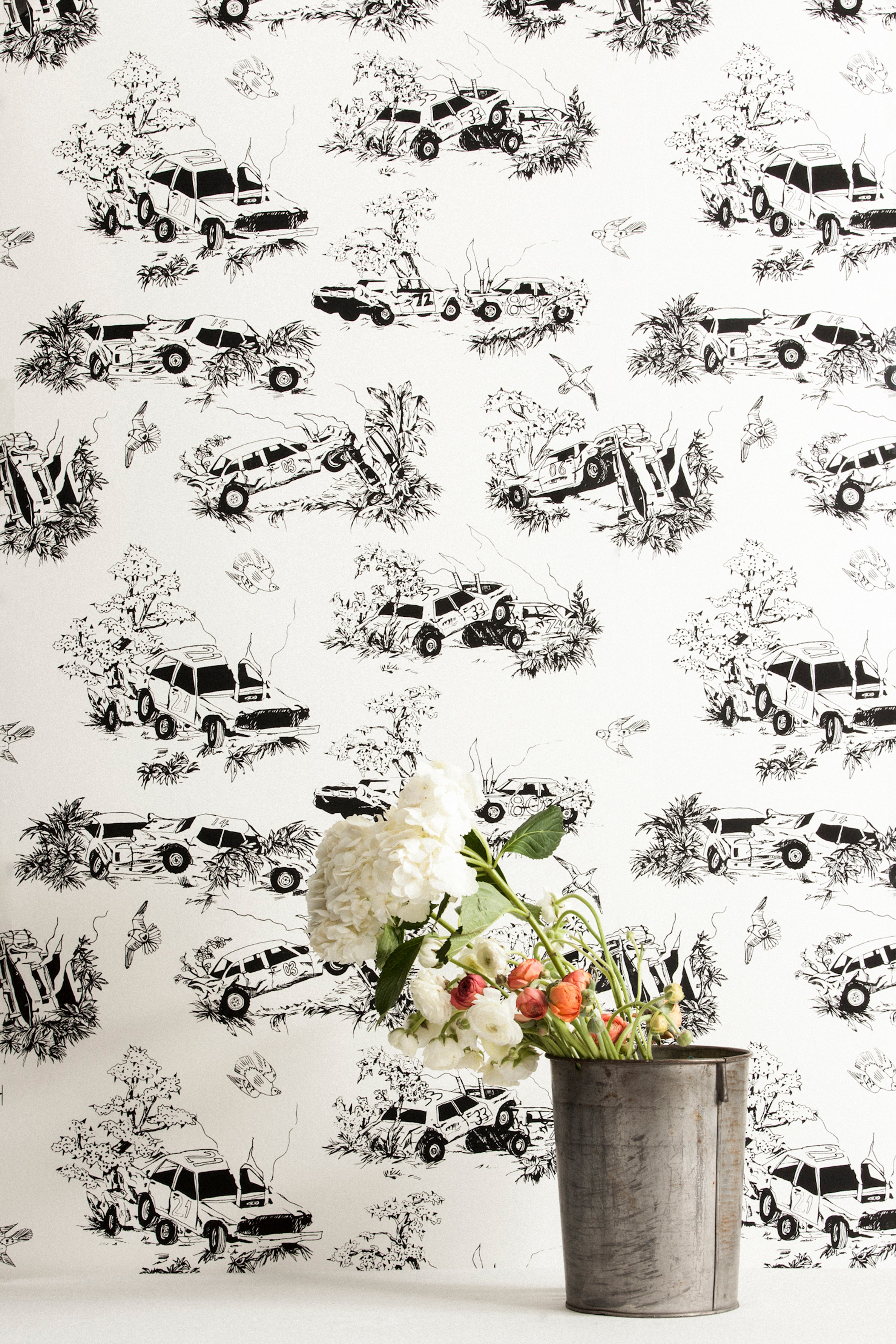 A vase of flowers stands in front of a wall covered in a playful illustrated car crash print in black on a white field.
