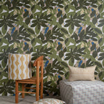 Several bolsters and a lamp sit in front of a wall papered in a botanical and parrot print in green, blue and yellow.