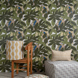 Several bolsters and a lamp sit in front of a wall papered in a botanical and parrot print in green, blue and yellow.