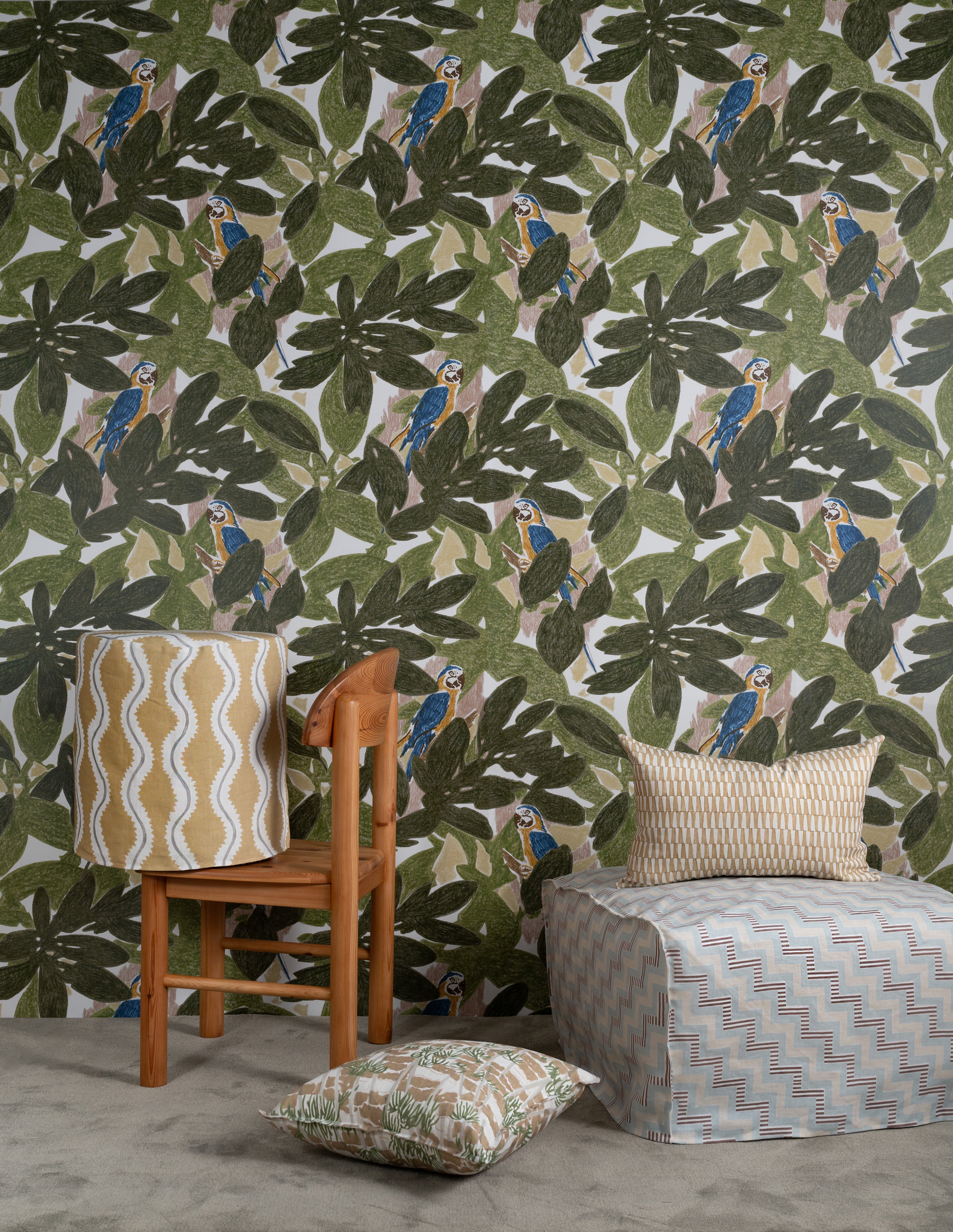 Several bolsters and a lamp sit in front of a wall papered in a botanical and parrot print in green, blue and yellow.