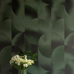 A vase of flowers stands in front of a wall covered in an abstract curvilinear print in mottled green and black.