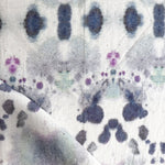 Draped fabric yardage in a watercolor inkblot print in shades of purple and gray on a cream field.