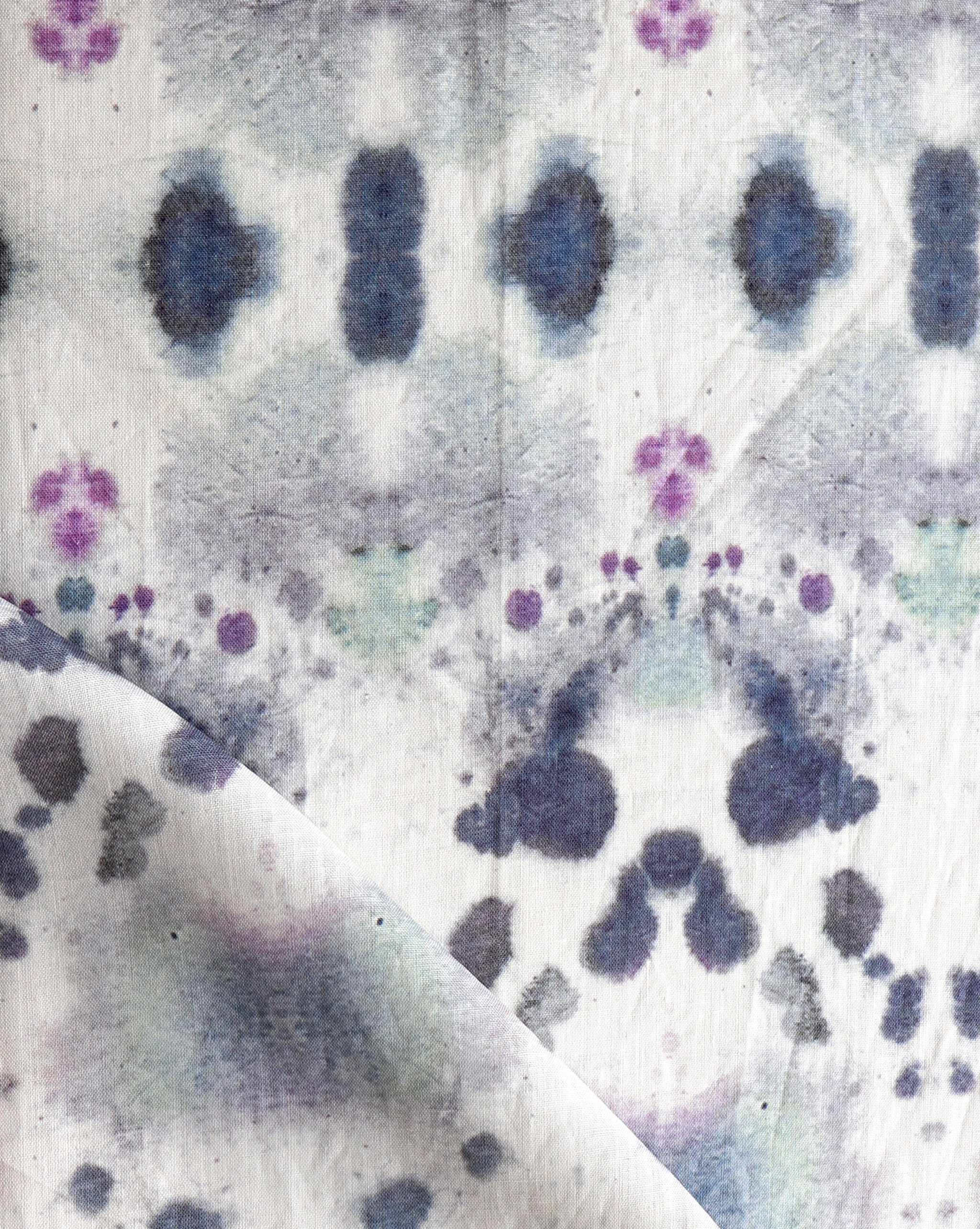 Draped fabric yardage in a watercolor inkblot print in shades of purple and gray on a cream field.