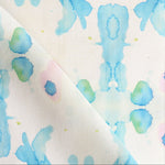 Draped fabric yardage in a watercolor ink splash print in shades of blue and green on a cream field.