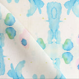Draped fabric yardage in a watercolor ink splash print in shades of blue and green on a cream field.