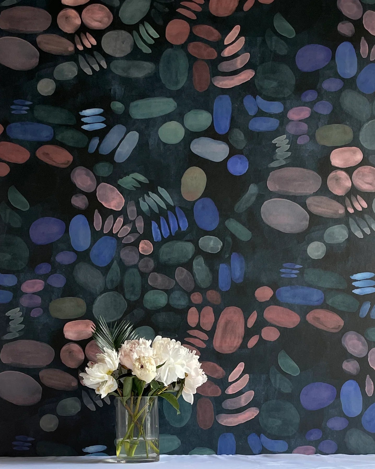 A vase of flowers stands in front of a wall covered in a playful paint blotch print in blue, green and pink on a black field.