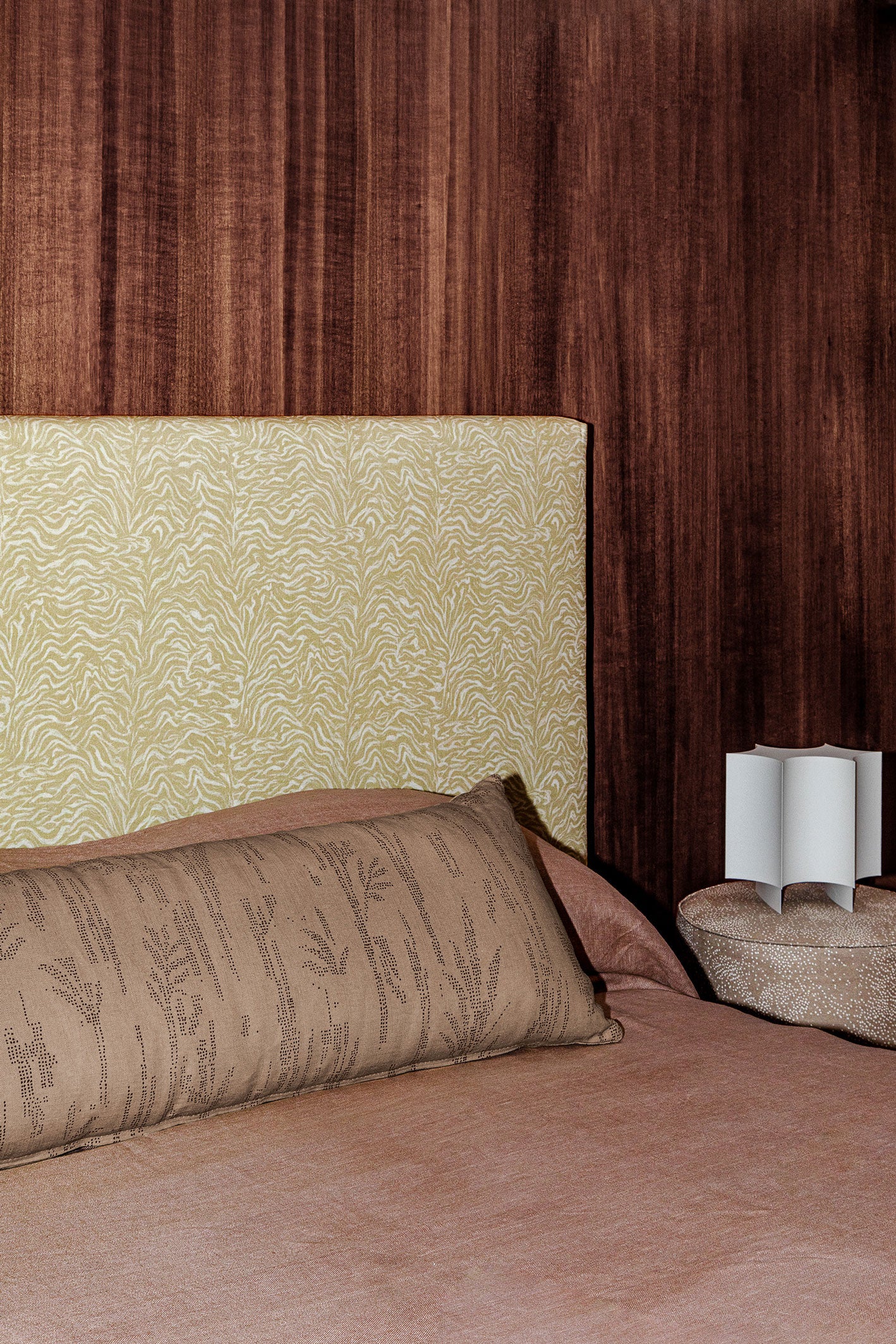 Bedroom scene with dark wood paneled wall, tan and white zebra headboard, clay colored bedding and an upholstered ottoman with white sculputral lamp