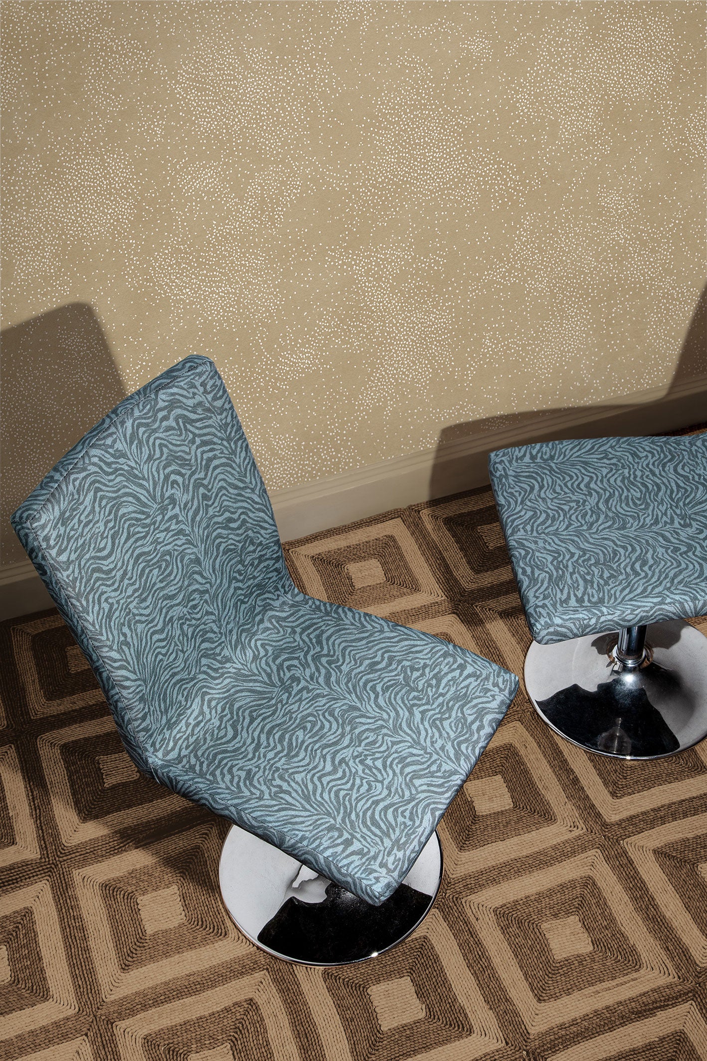 Overhead shot of two upholstered pestal chairs with a blue zebra pattern upholstery on a square patterned rug in brown against wallaper with white dots scattered on a tan field.