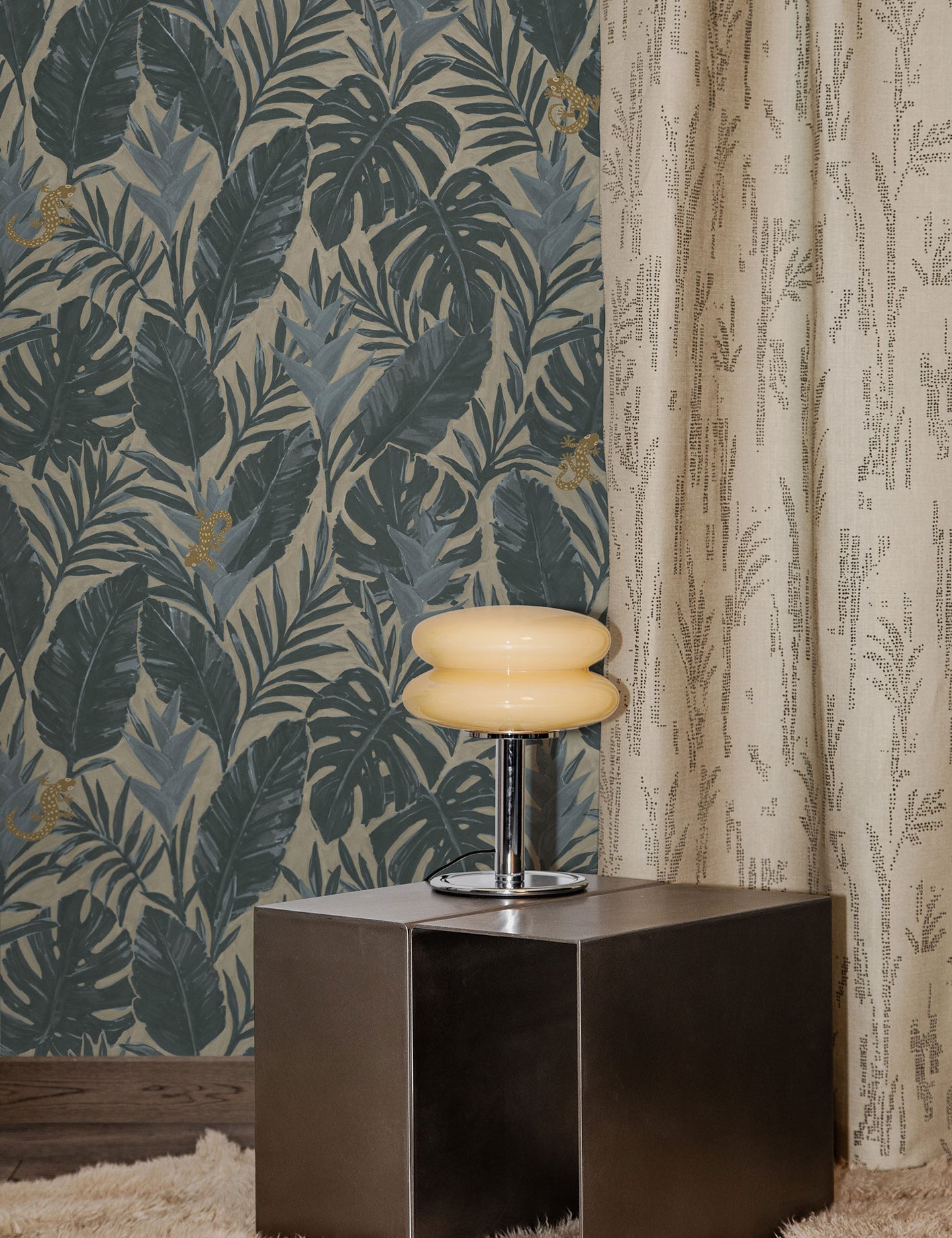 Styled interior with steel side table, retro chrome lamp with cream glass shade on a beige shag carpet against a teal and grey palm frond wallpaper and floor length curtain with a subtle dotted pattern in beige.