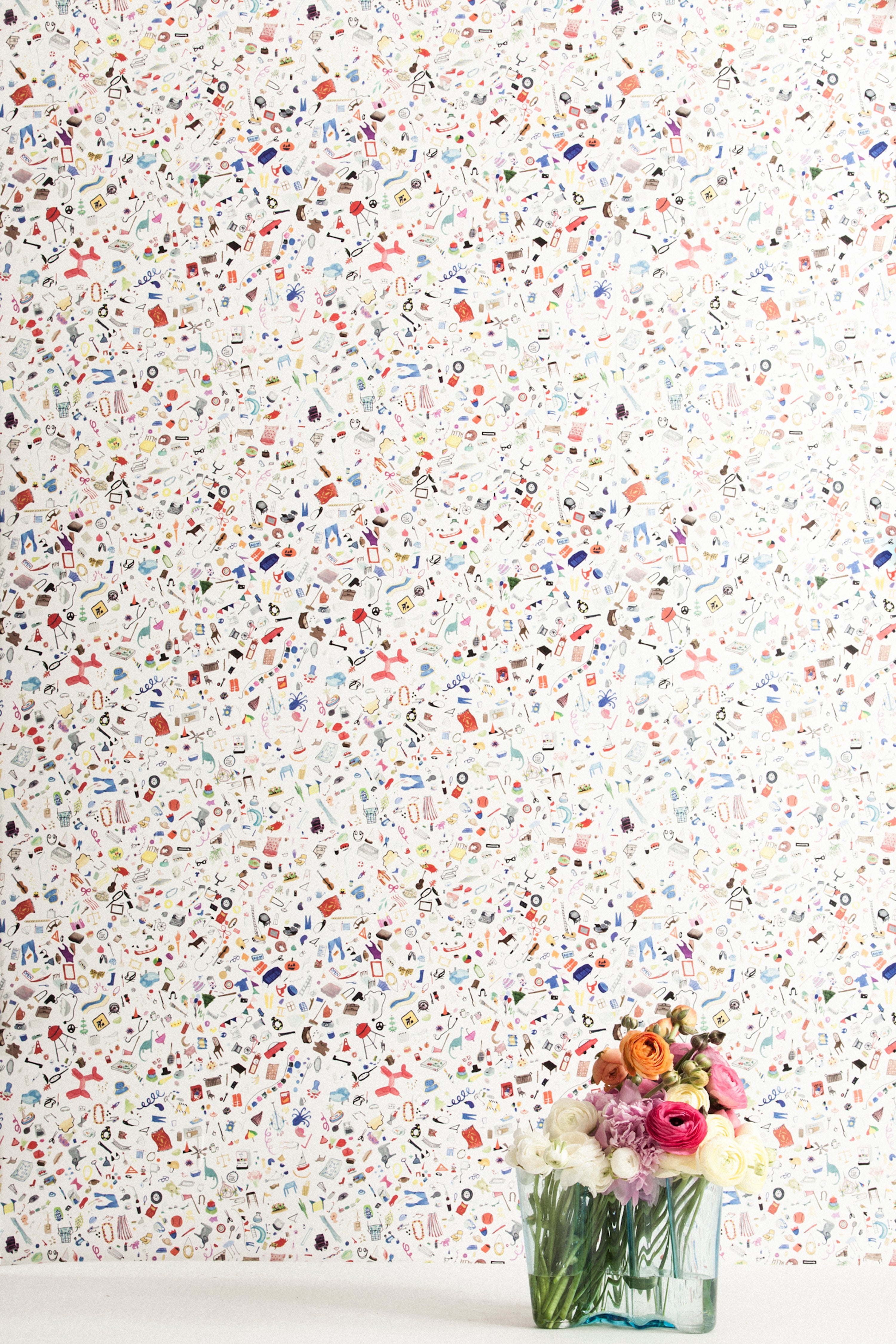 A vase of flowers stands in front of a wall papered in a playful illustrated "stuff" print in multicolor on a white field.
