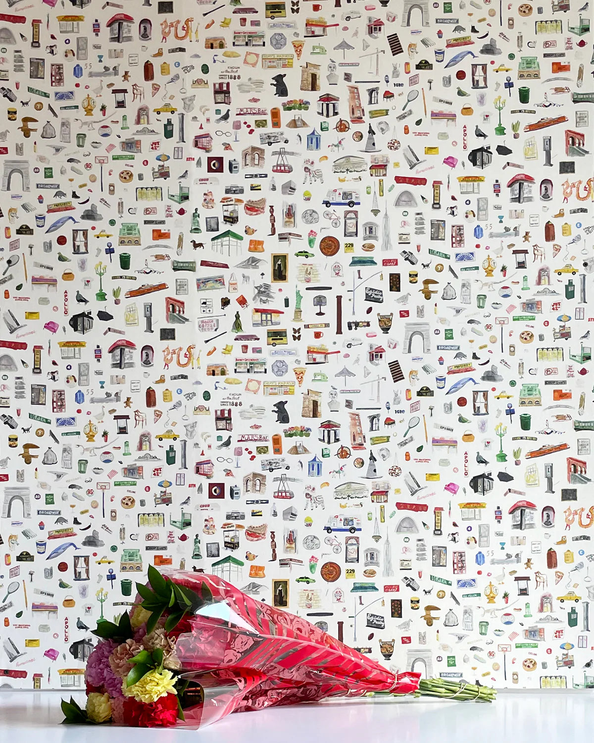 A bouquet of flowers lays in front of a wall papered in a playful illustrated "stuff" print in multicolor on a white field.