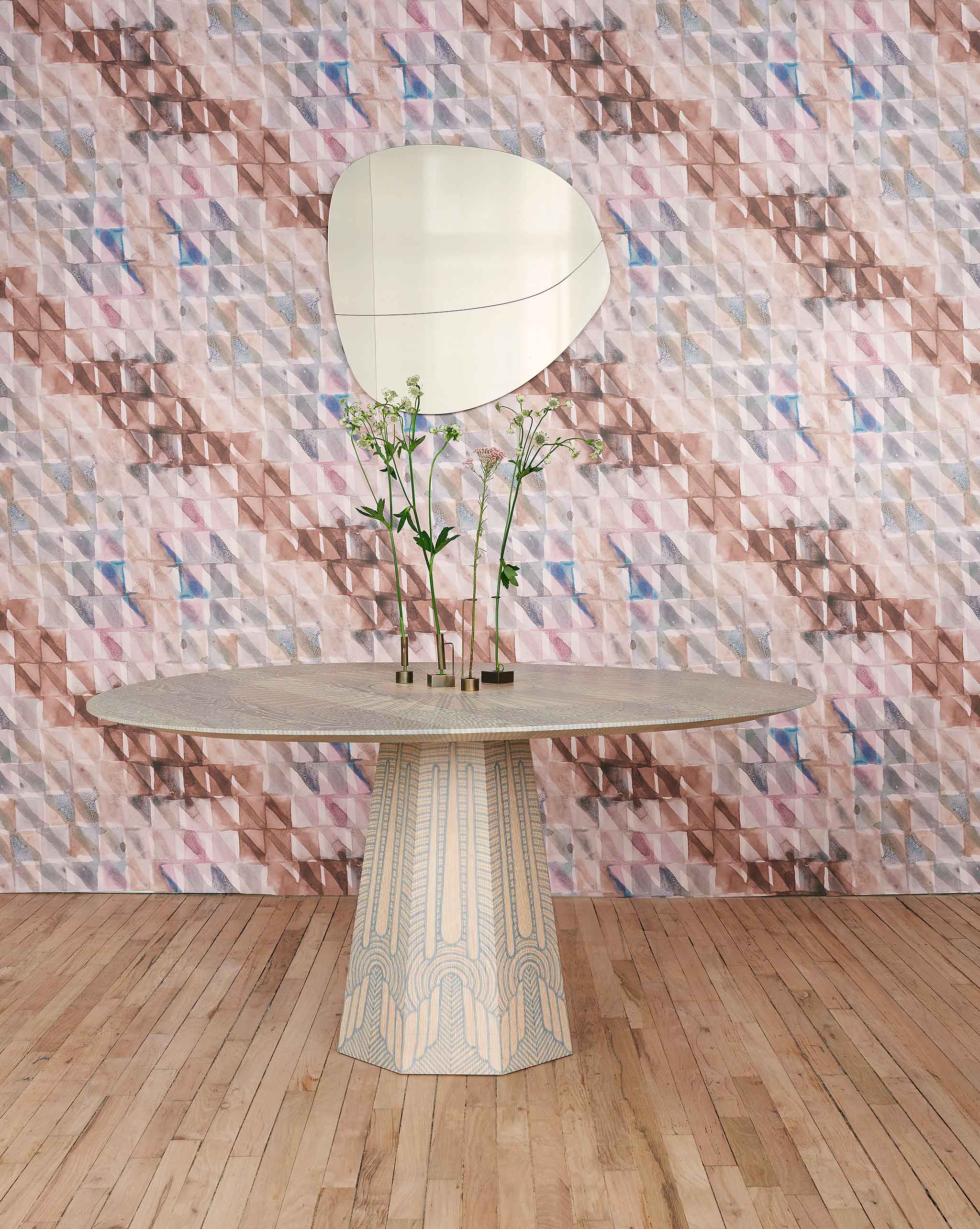 A modernist table with flowers stands in front of a wall papered in a painterly check pattern in brown, blue and pink.
