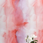 A vase of flowers stands in front of a wall papered in an abstract curvilinear pattern in mottled pink, red and purple.