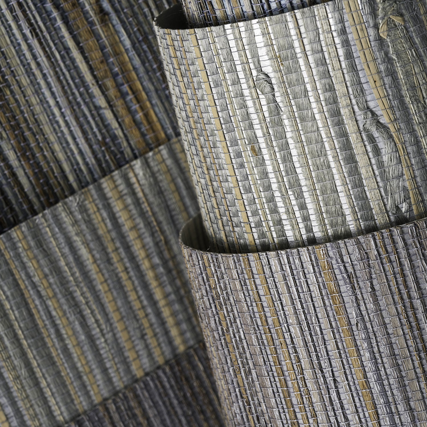 A trio of rolls of interior fabrics, all in shades of metallic silver, standing in front of a wall covered in another striped silver fabric.