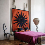 Bedroom scene with a dramatic sculptural chair, pedestal with urn, and a magenta bedspread against a rug tapestry with a bold sun motif in black on red. 