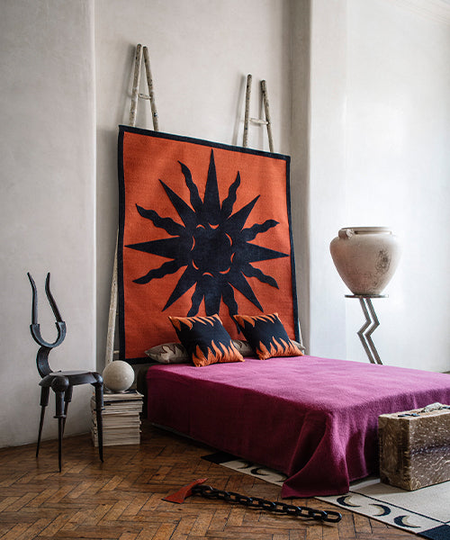 Bedroom scene with a dramatic sculptural chair, pedestal with urn, and a magenta bedspread against a rug tapestry with a bold sun motif in black on red. 