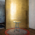 Golden curved room designer and verdigris side table against handknotted circular rug with a flame border design in red on a brown field. 