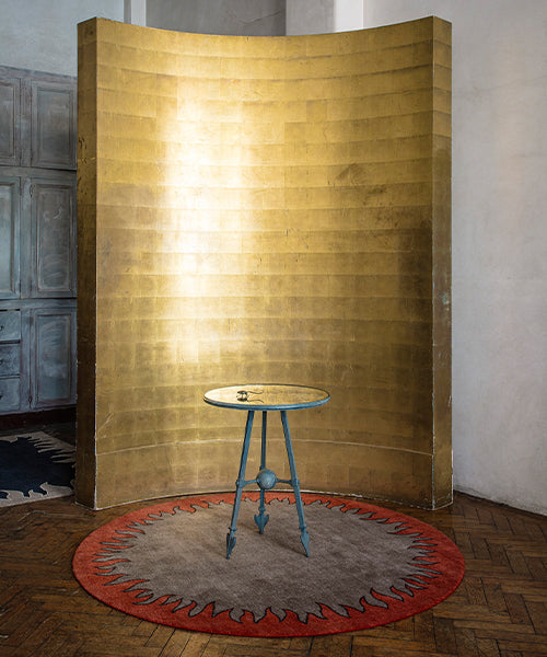 Golden curved room designer and verdigris side table against handknotted circular rug with a flame border design in red on a brown field. 