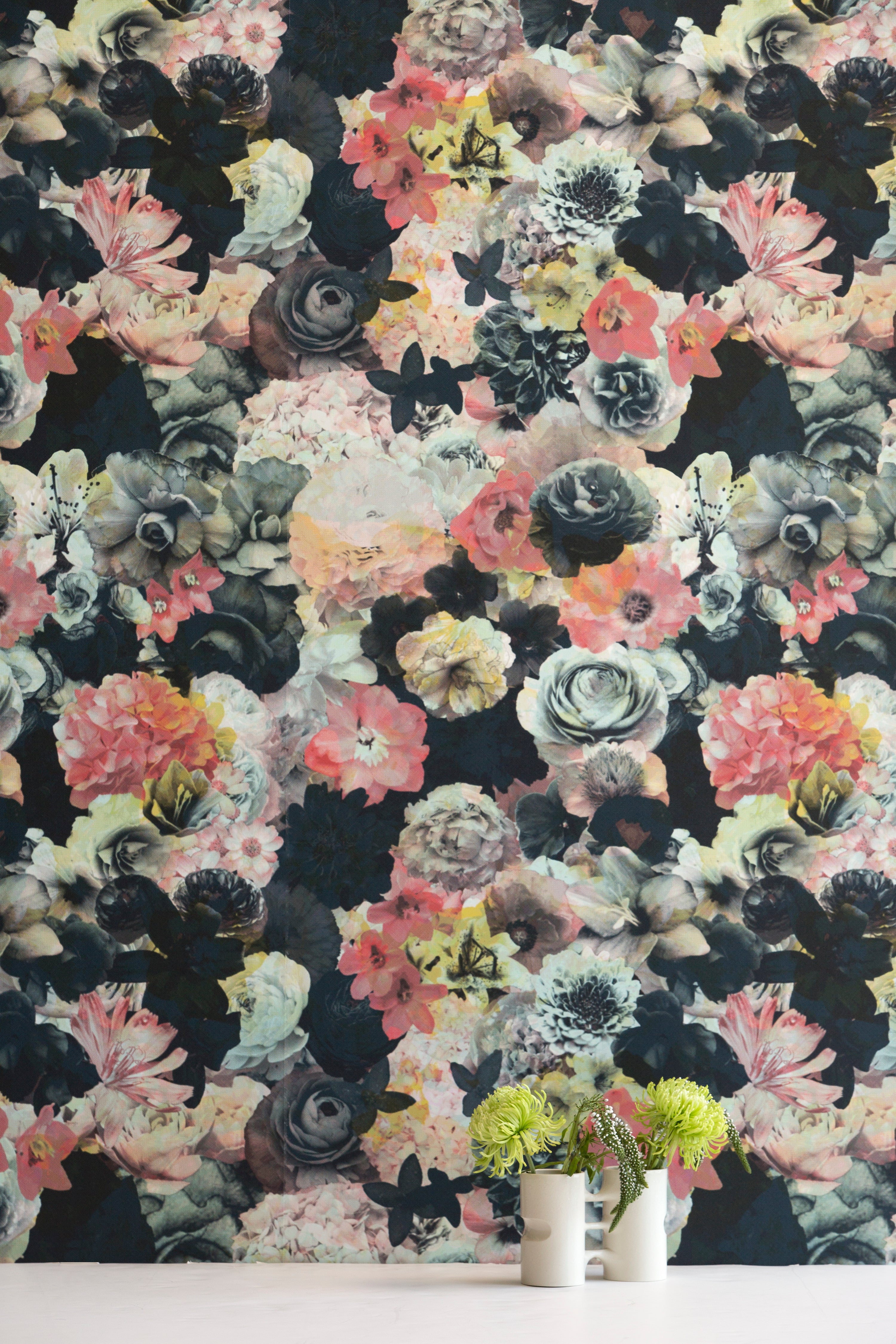 A vase of flowers stands in front of a wall covered in a dense photorealistic floral print in pink and green on a navy field.