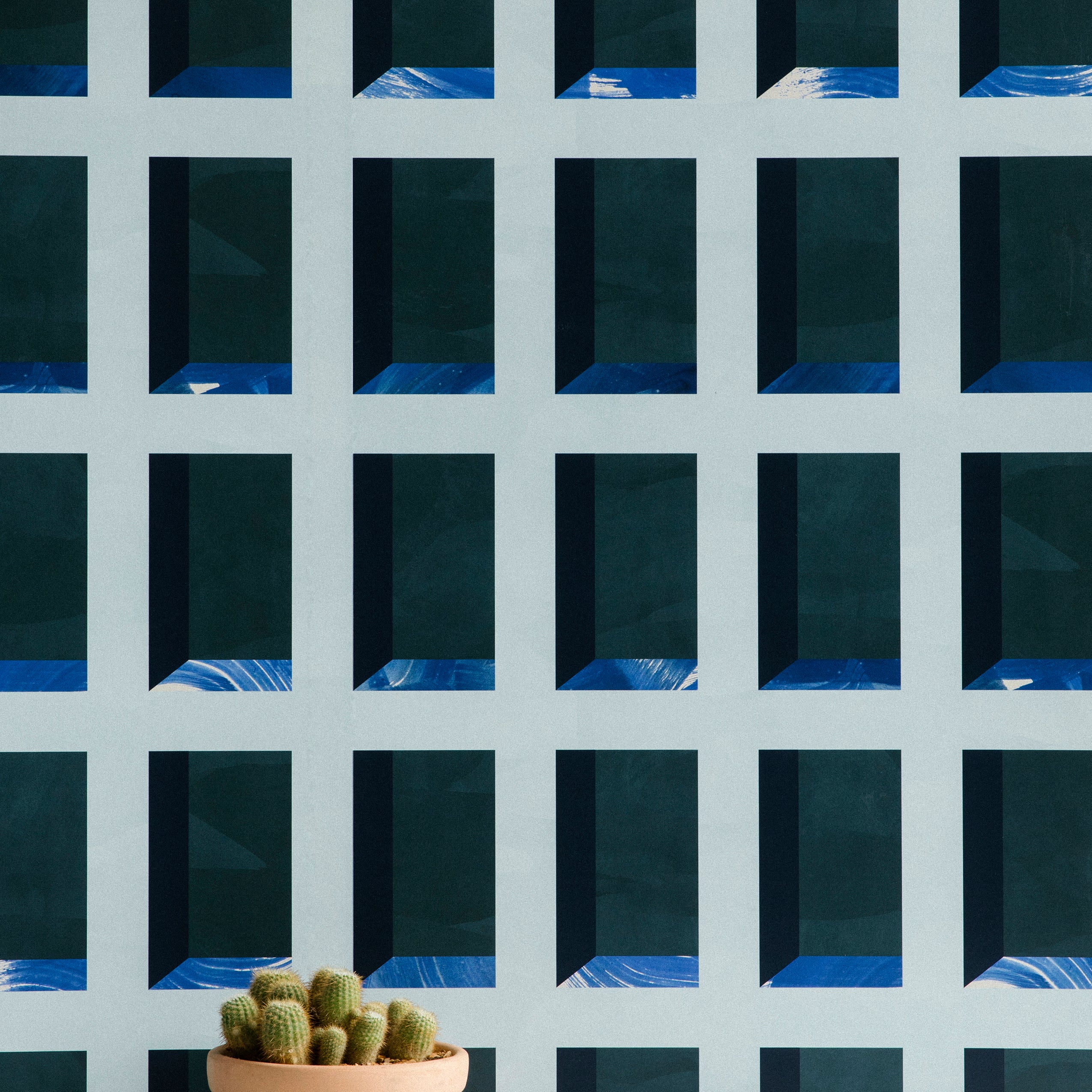 A potted cactus stands in front of a wall papered in a dimensional grid print in shades of blue, navy and turquoise.