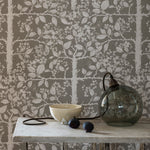 Cluttered end table in front of an accent wall papered in a large-scale tree and leaf print in white on a light gray field.
