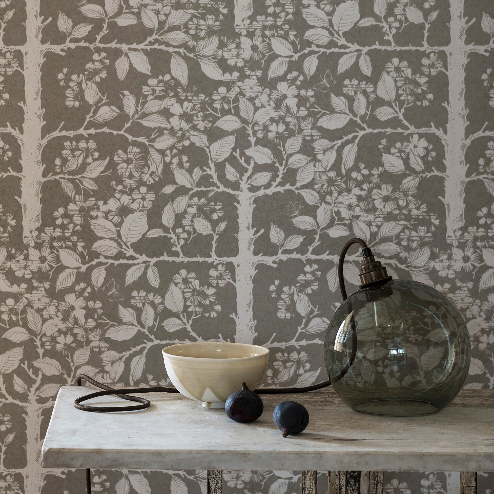 Cluttered end table in front of an accent wall papered in a large-scale tree and leaf print in white on a light gray field.