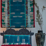 Ornamental wooden bench with bod watercolor cushions in turquoise against a hanging panel in the same design.