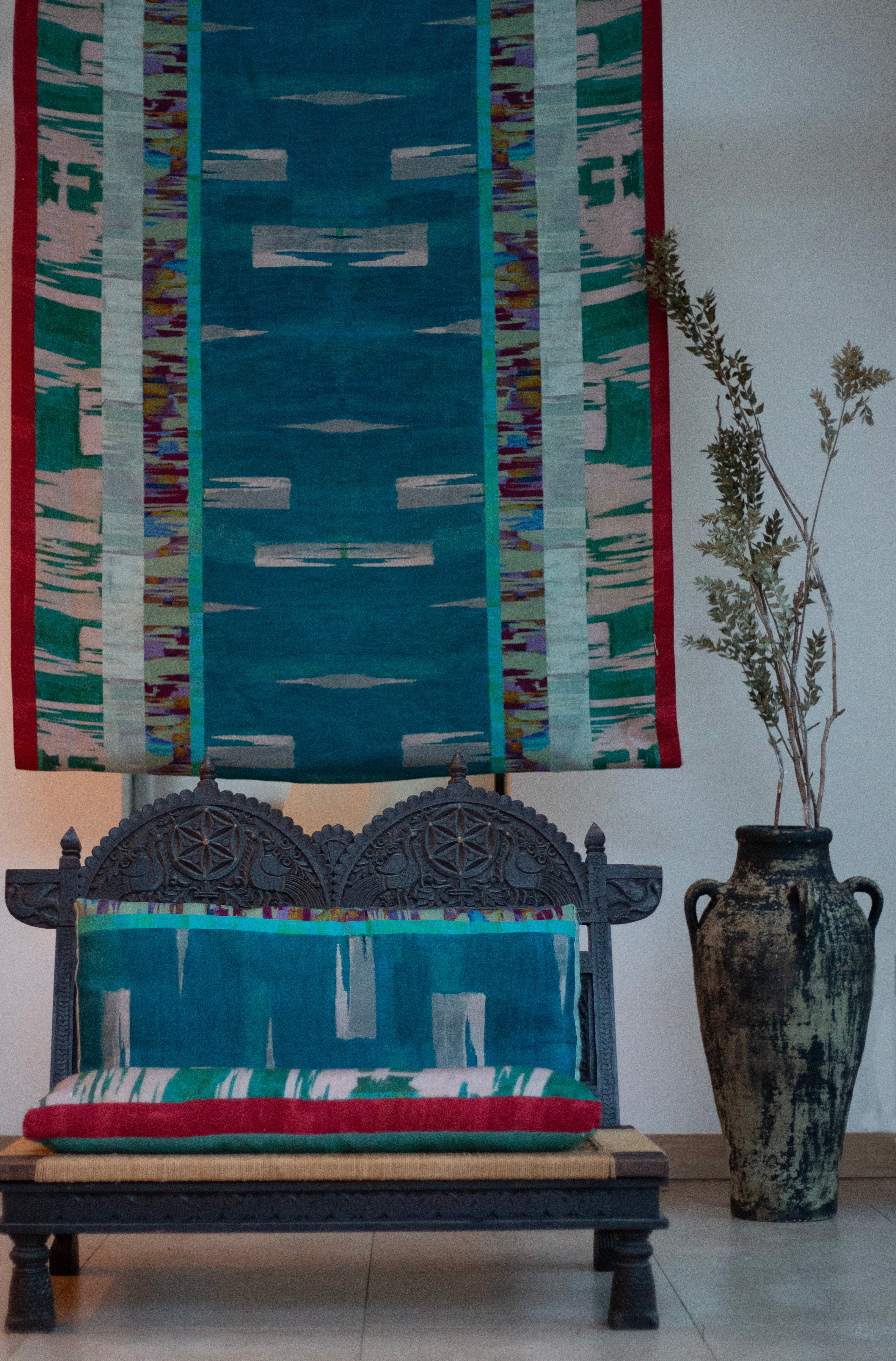 Ornamental wooden bench with bod watercolor cushions in turquoise against a hanging panel in the same design.