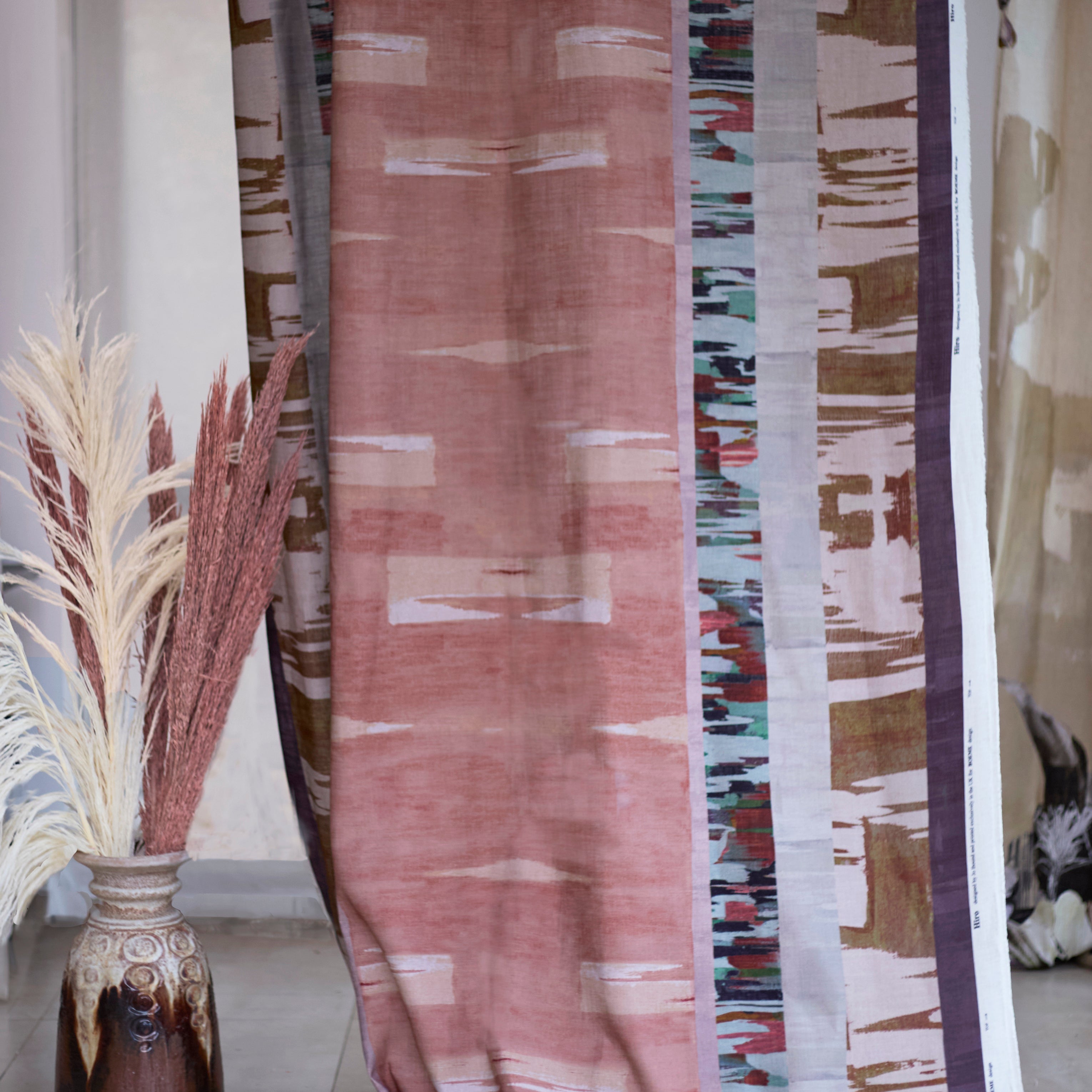 Curtain in a Printed fabric in an engineered stripe design with watercolor effects in shades of blush, plum, brown and aqua, with a ceramic urn with dried grasses.