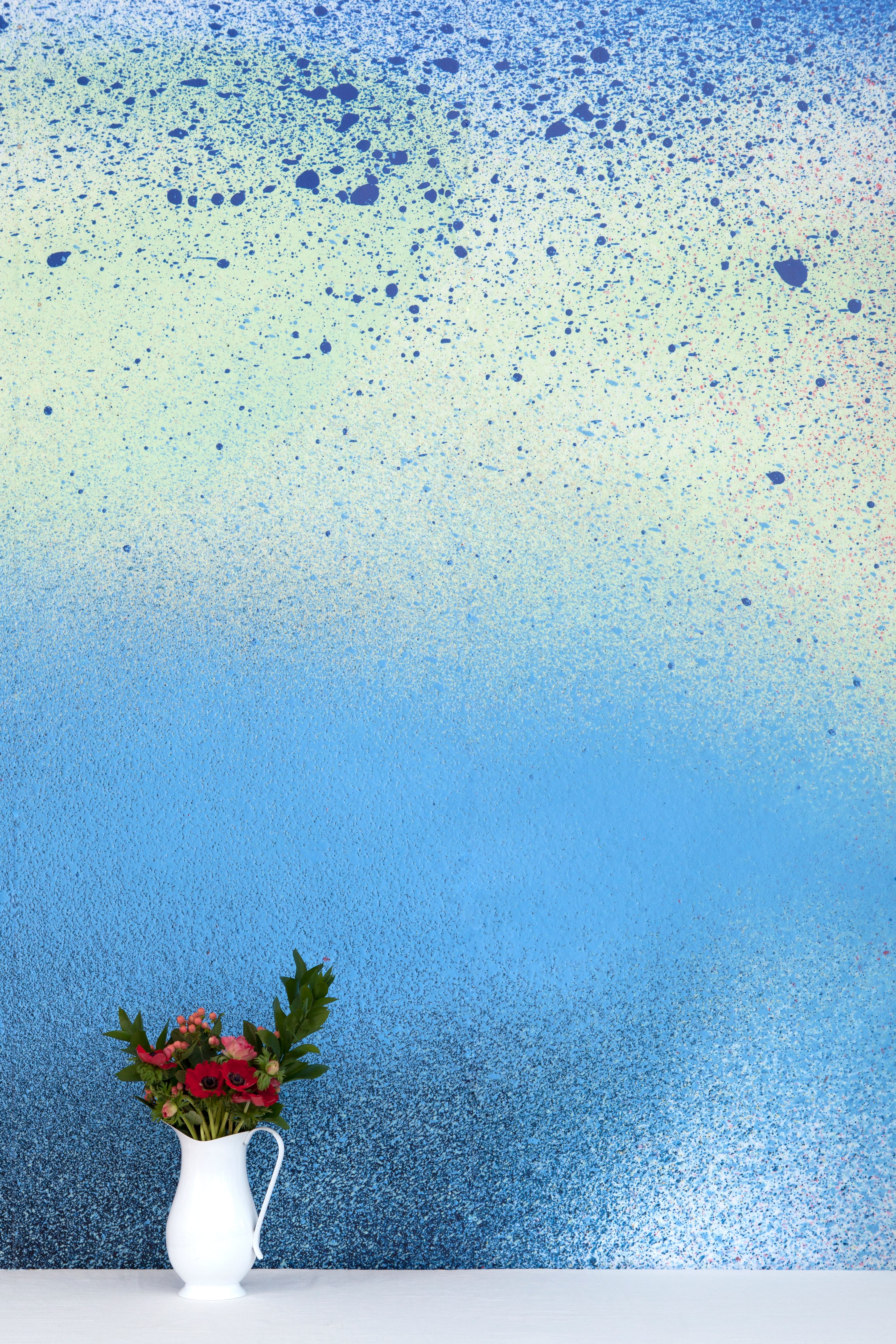 A vase of flowers stands in front of a wall covered in an abstract ombré paint splatter print in blue, navy and green.