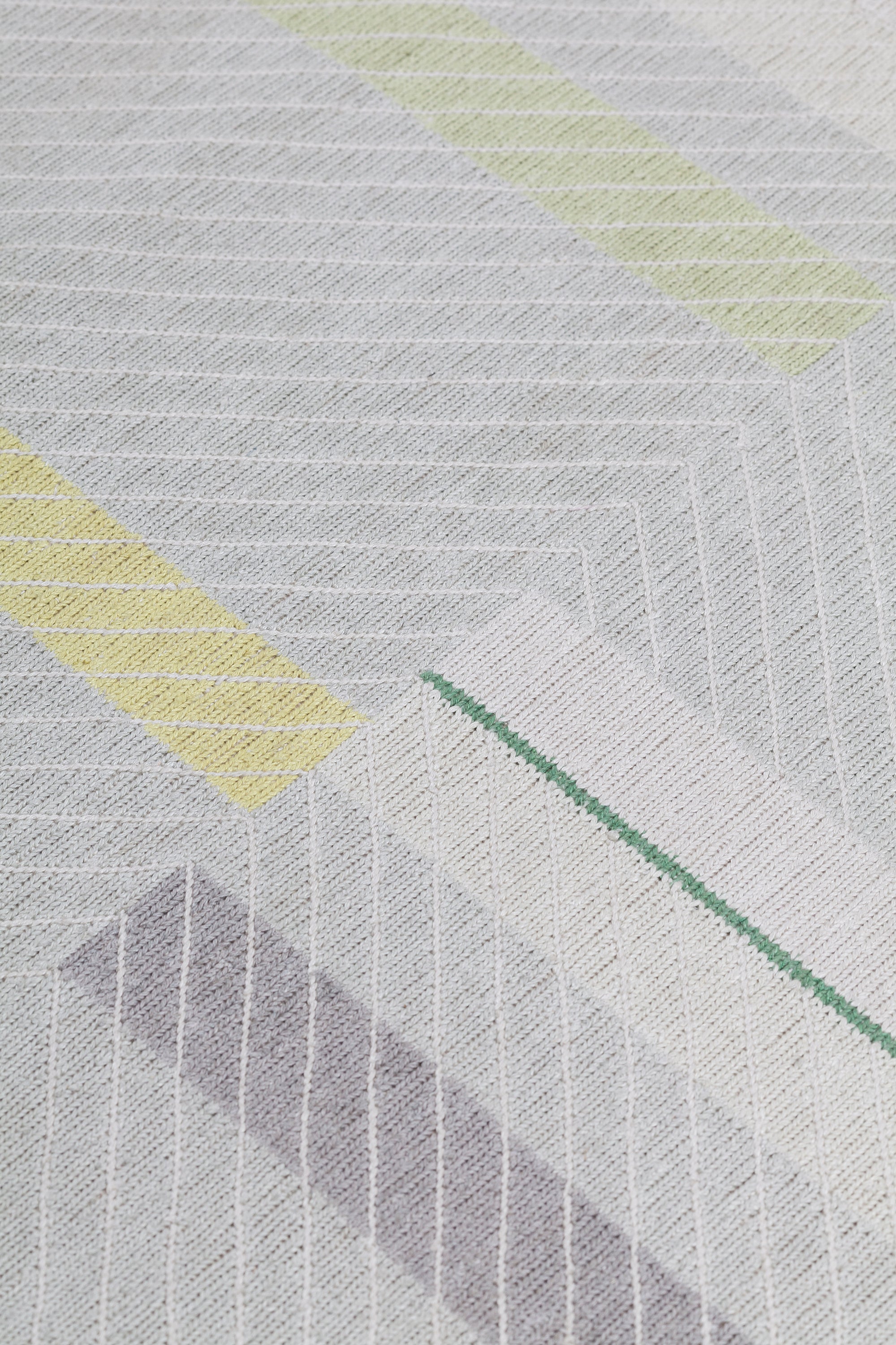 Detail of the Amelia Rug in Peridot featuring a minimalist broken stripe pattern, overlayed with thin white diagonal lines. The broken stripes are a mix of white, grey, and shades of yellow with kelly green accents, all on a light grey field. 