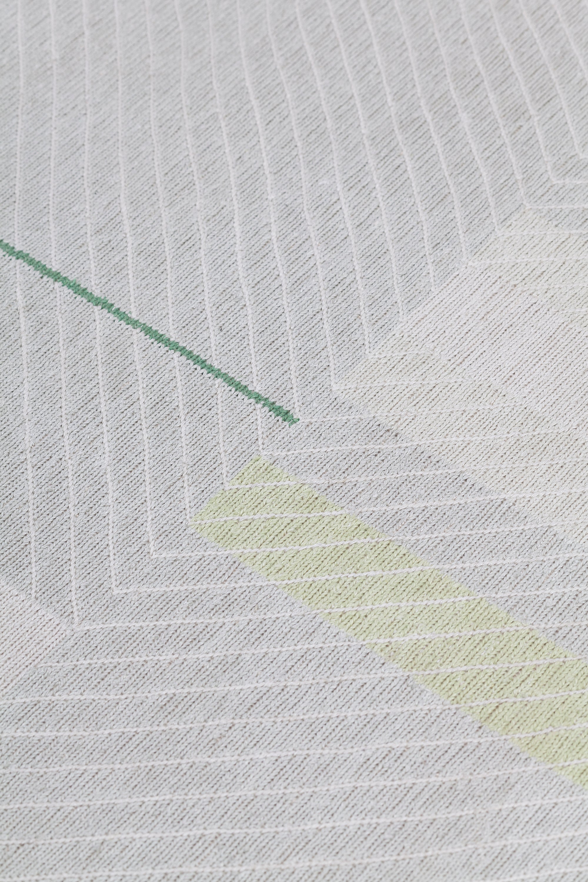 Detail of the Amelia Rug in Peridot featuring a minimalist broken stripe pattern, overlayed with thin white diagonal lines. The broken stripes are a mix of white, grey, and shades of yellow with kelly green accents, all on a light grey field. 