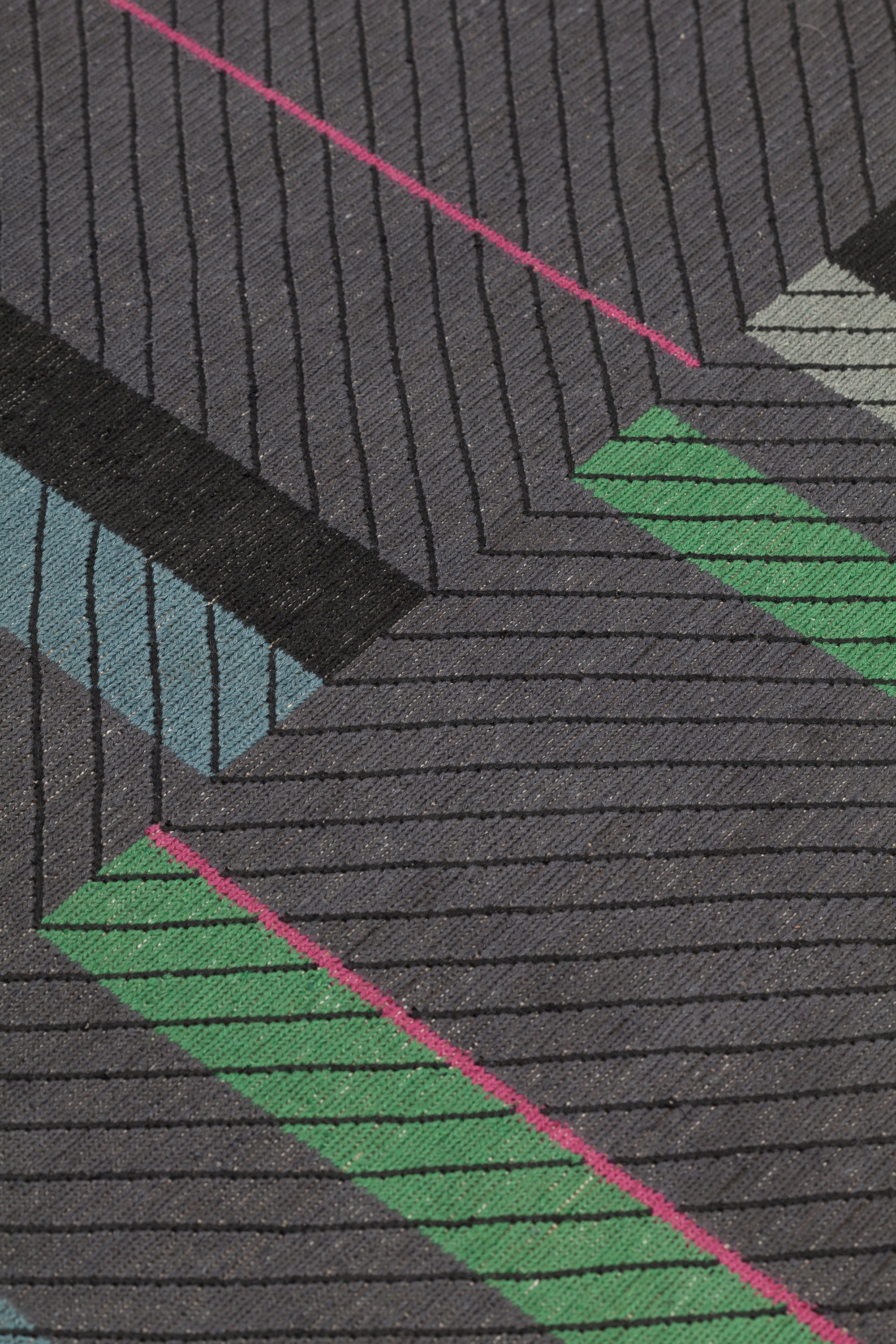 Detail of the Amelia Rug in Spinel featuring a minimalist broken stripe pattern, overlayed with thin black diagonal lines. The broken stripes are a mix of white, grey, kelly green, denim blue, black with hot pink accents, all on a charcoal grey field. 