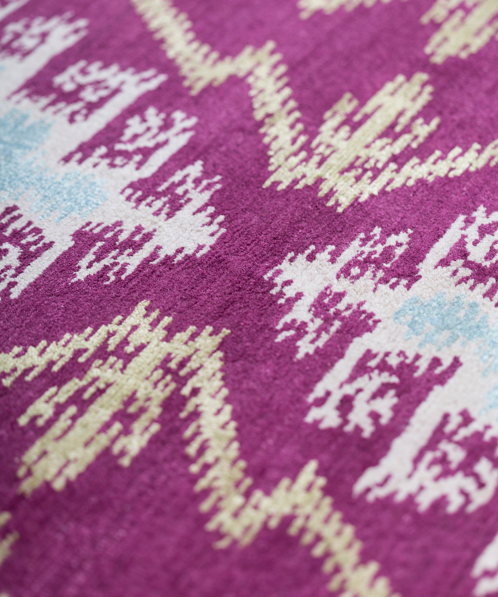 Detail of the Anna Ikat rug in Cranberry features an ikat inspired pattern of diamonds in blue and white with pale lime green accents on a magenta field