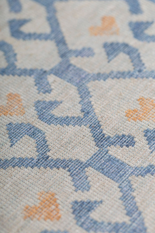 Detail of the Avesta Rug in Blue, a blue lattice pattern on a beige field with small orange heart accents. 