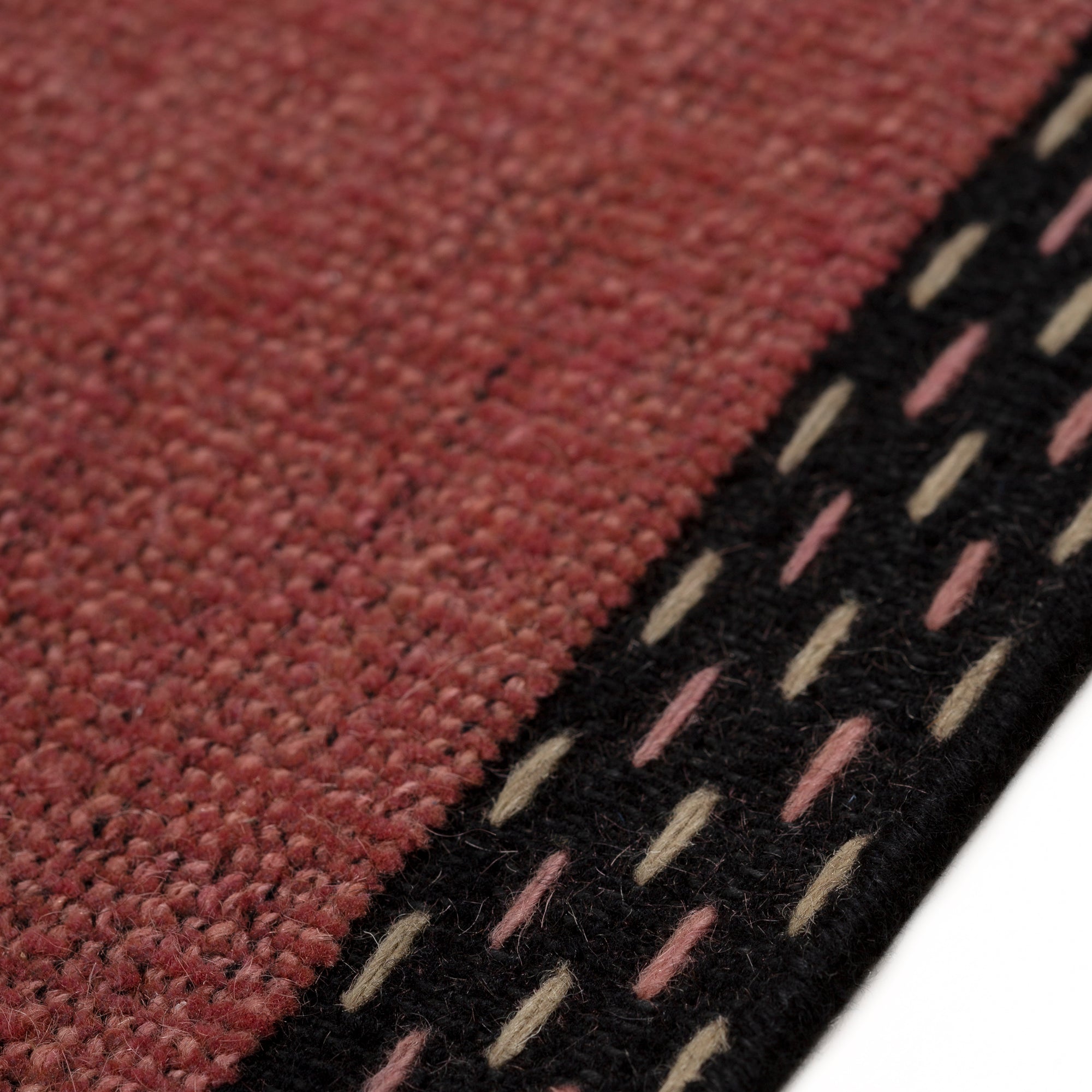 Detail of the Plain Stitched Border Rug in Carmine, a solid red rug with a black border with black border with pink and taupe dashes. 