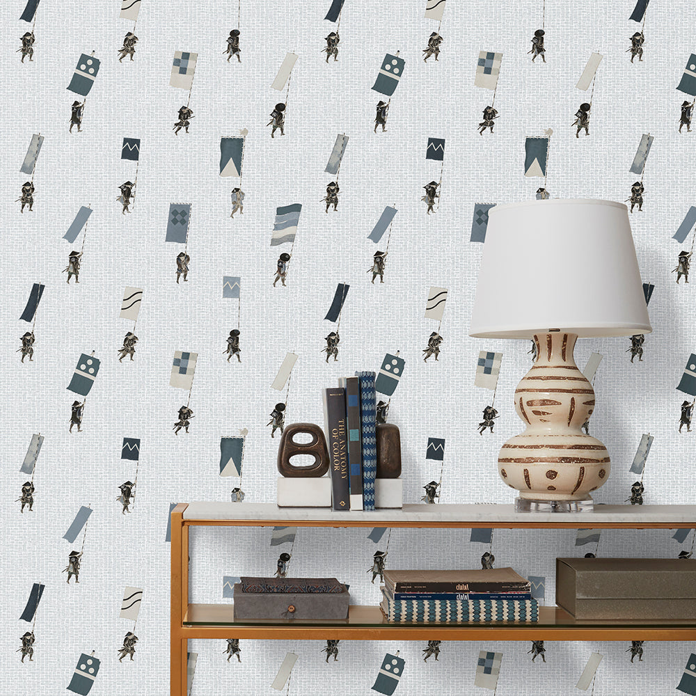 A table with books and a lamp in front of a wall papered in a pattern of Japanese soldiers carrying flags on a light gray background.
