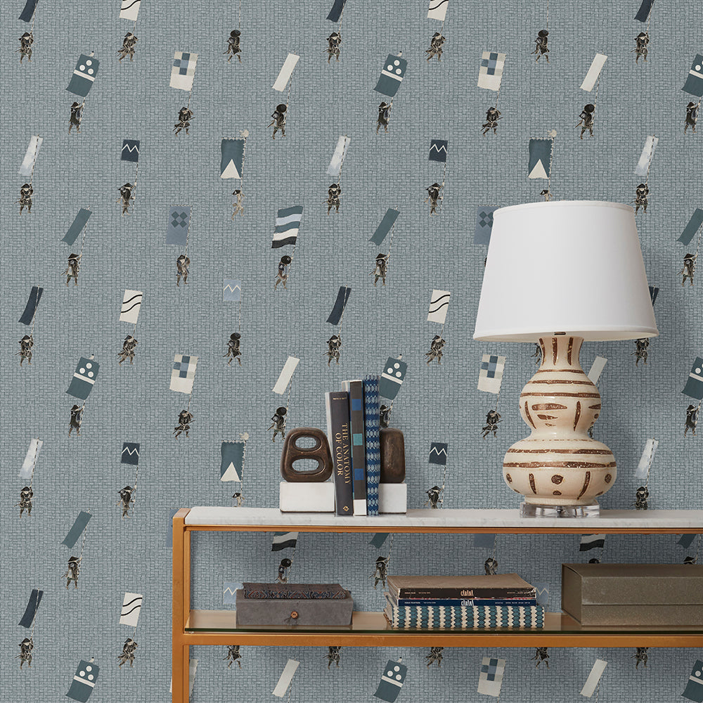 A table with books and a lamp in front of a wall papered in a pattern of Japanese soldiers carrying flags on a blue-gray background.